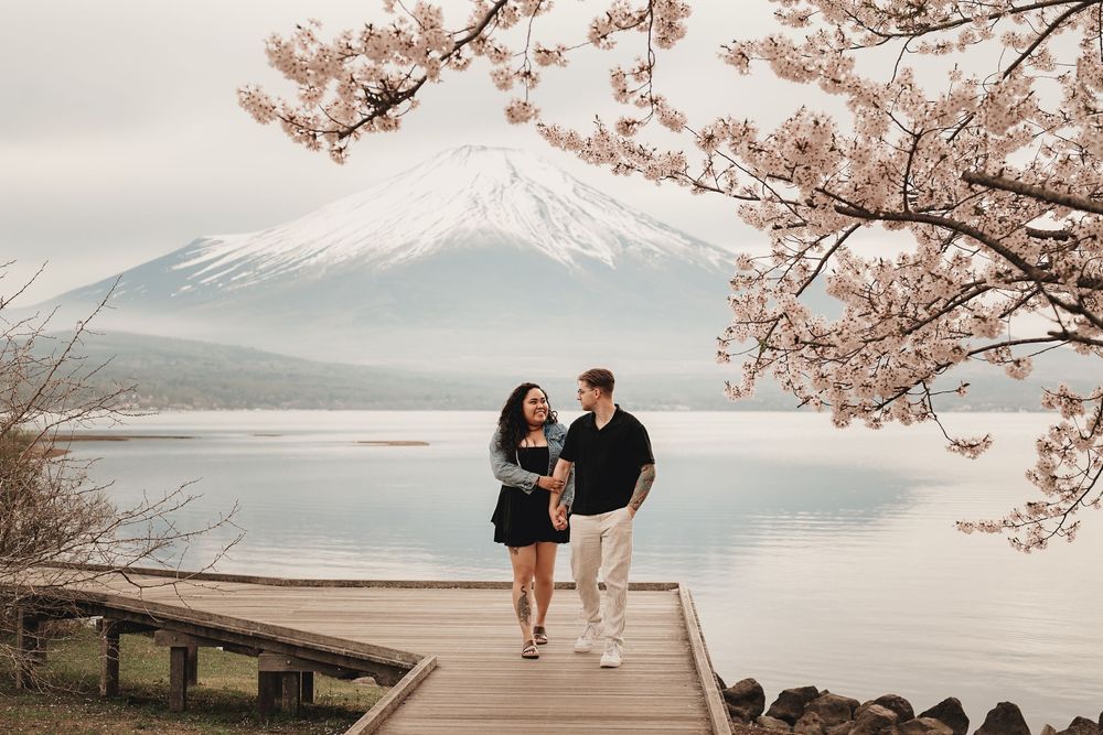 A couple stands on a wooden pier by Lake Kawaguchiko with Mount Fuji and cherry blossoms in the background.