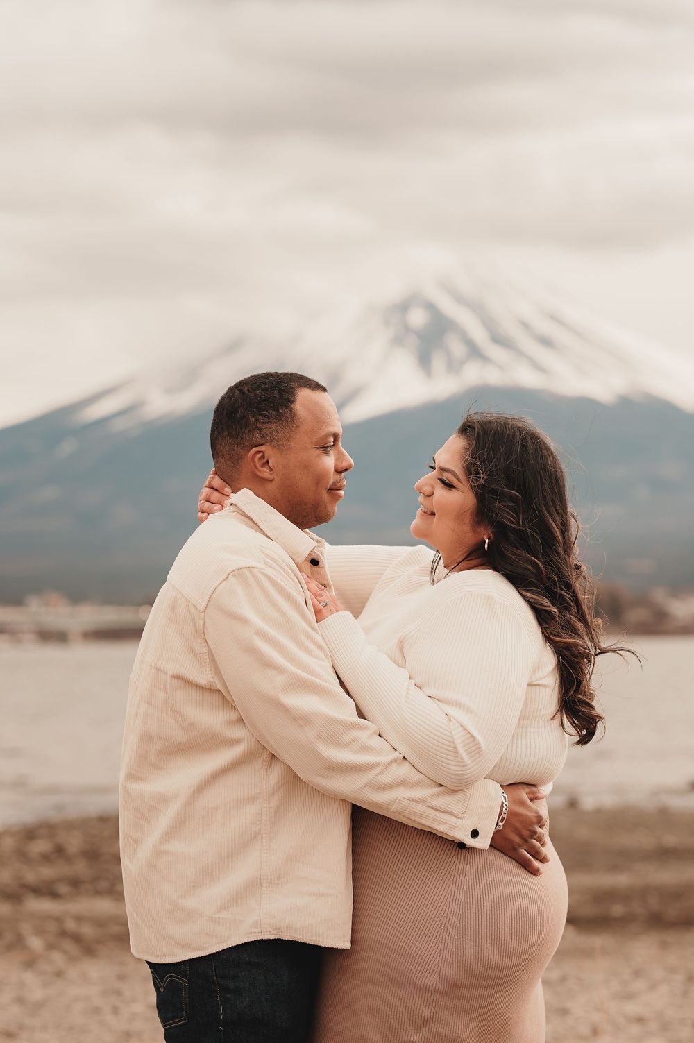 A loving couple embraces in neutral colored outfits while standing near Mount Fuji in Japan.