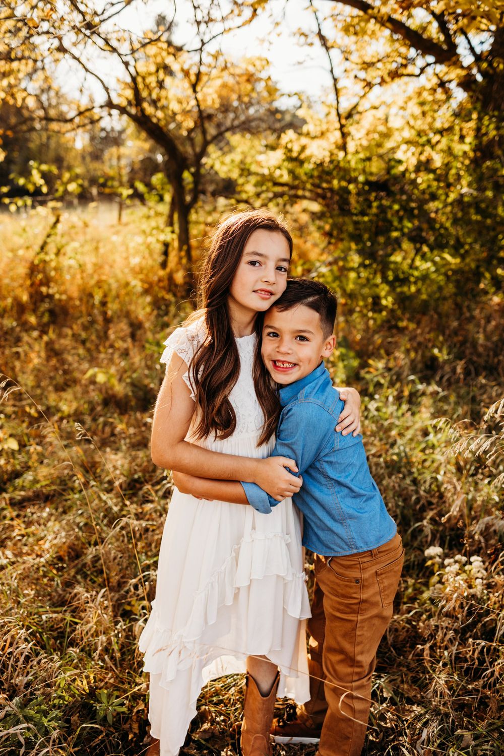 Two children share a sweet embrace in a golden autumn setting with sunlit trees in the background.