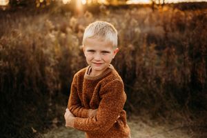 Child wearing brown sweater standing in tall grass during golden hour sunset.