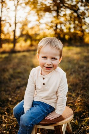 A child sits on a wooden stool outdoors during sunset with golden light creating a warm autumn atmosphere.