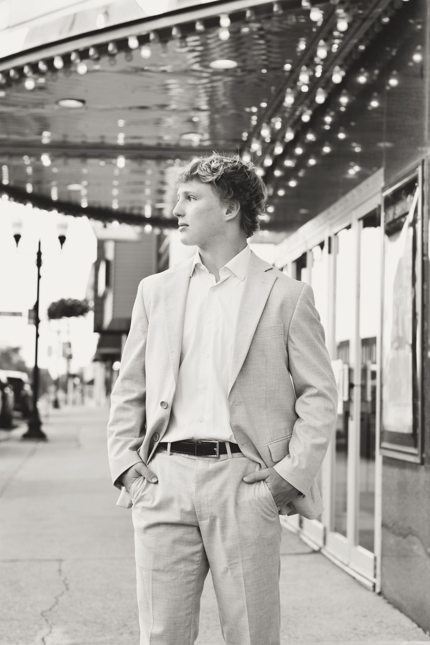 Black and white portrait of senior in light suit standing under string lights.