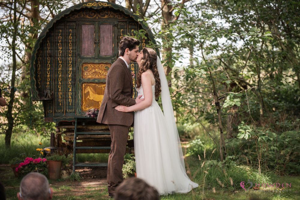 A romantic vintage gypsy wagon serves as a backdrop for a couple sharing a wedding moment in a woodland setting.