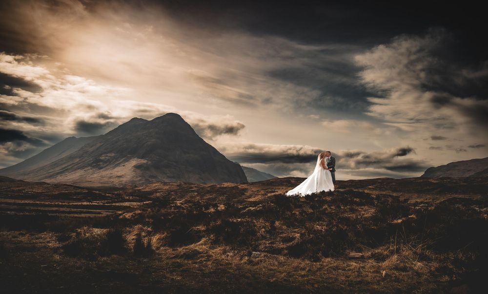 Dramatic wedding photo silhouette against a mountainous Scottish Highland landscape at sunset with moody clouds.