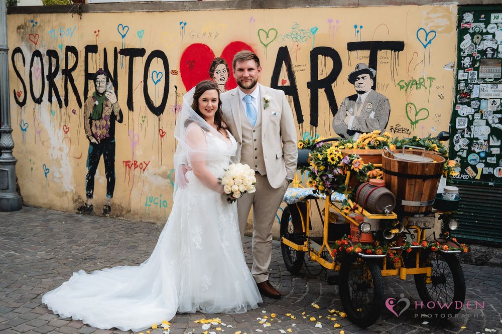 Wedding couple poses by graffiti wall in Sorrento, Italy with antique bike and floral bouquet against urban backdrop.
