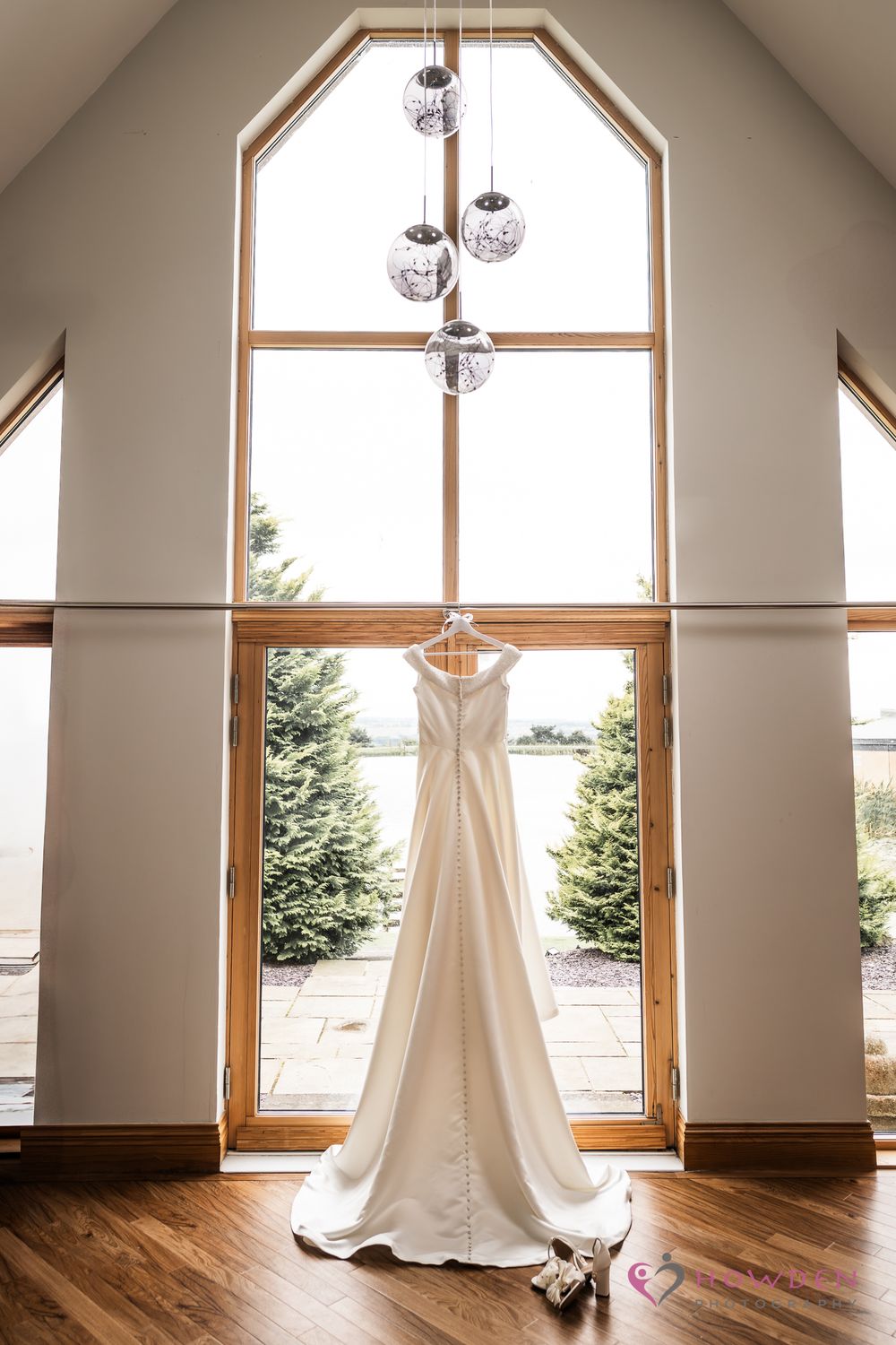 Wedding dress hangs elegantly in front of tall cathedral window with glass pendant lights and hardwood floors.