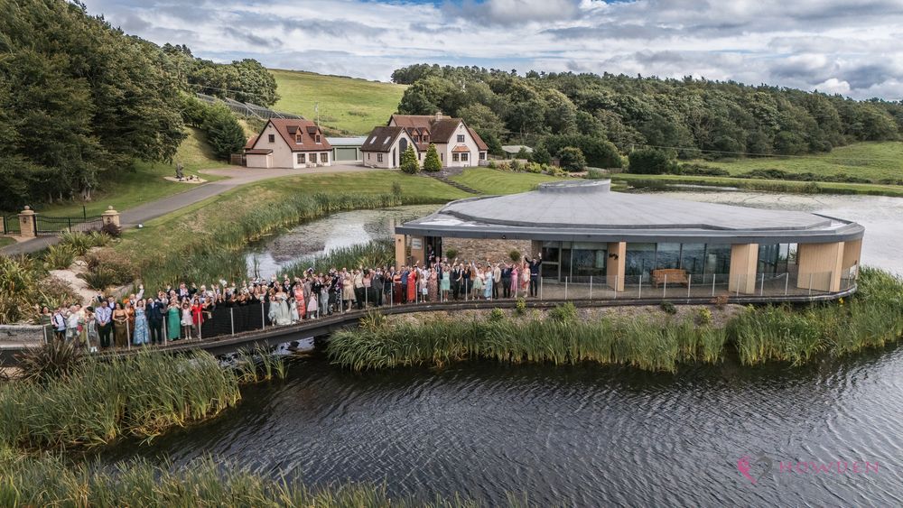 Aerial view of a modern visitor center building with curved green roof next to a lake and traditional cottages in Scotland.