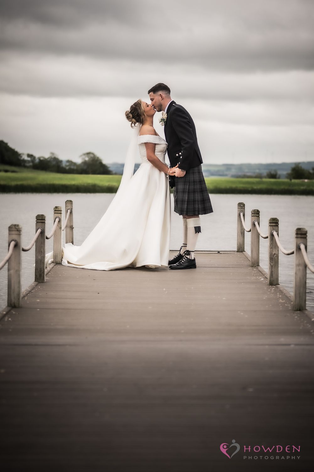 A romantic wedding couple share an intimate kiss on a wooden dock over a lake, the groom wearing a traditional Scottish kilt.