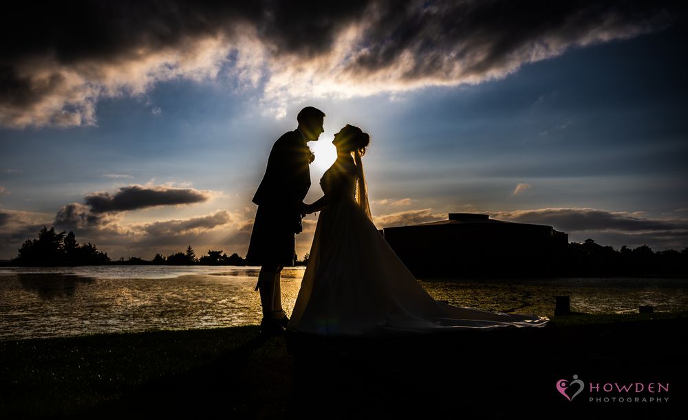 Silhouetted couple stands together against dramatic sunset sky with dark clouds in romantic wedding photo.