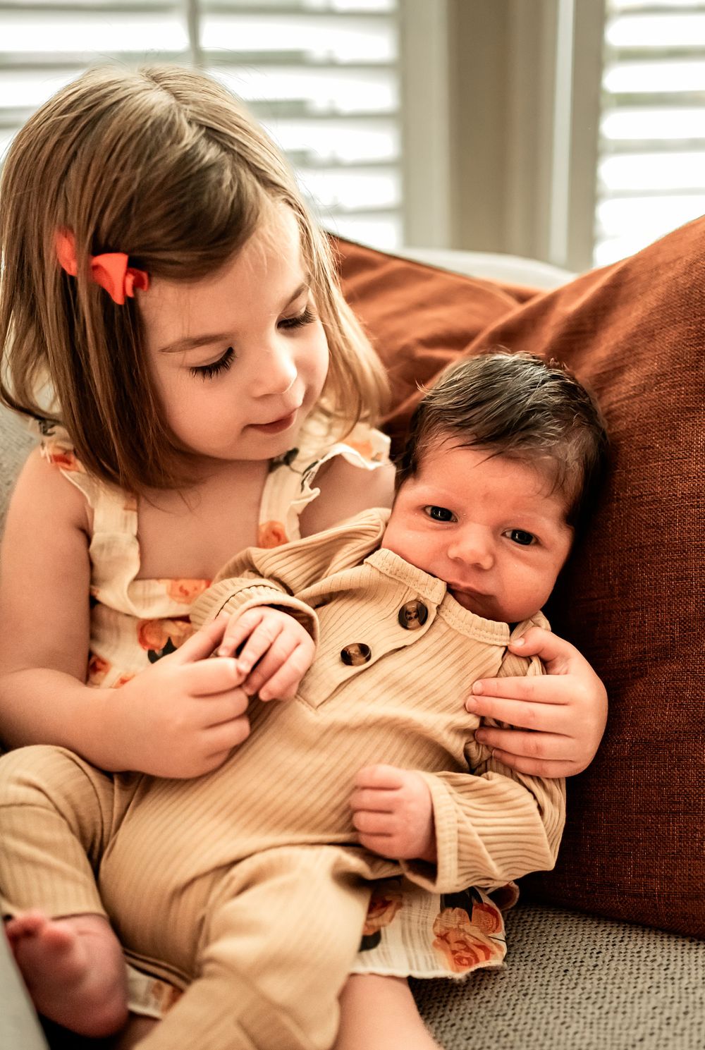 Siblings share a tender moment on a brown couch as the older sister holds the baby wearing a beige romper.