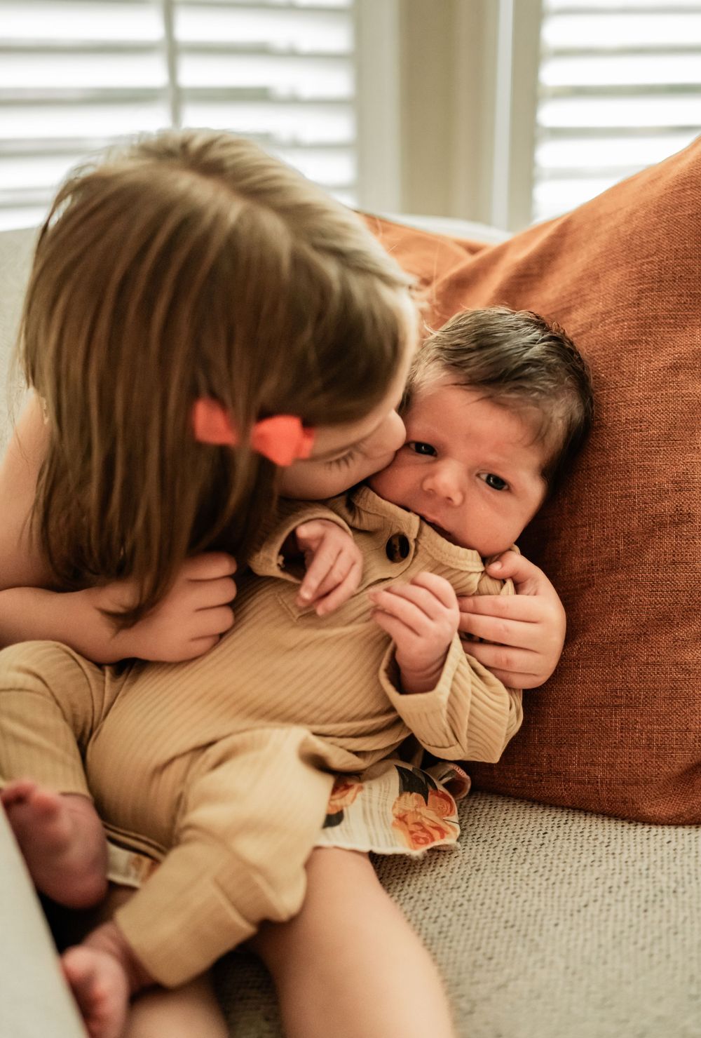 Sweet sibling interaction with older sister snuggling baby in beige outfit against orange pillow.