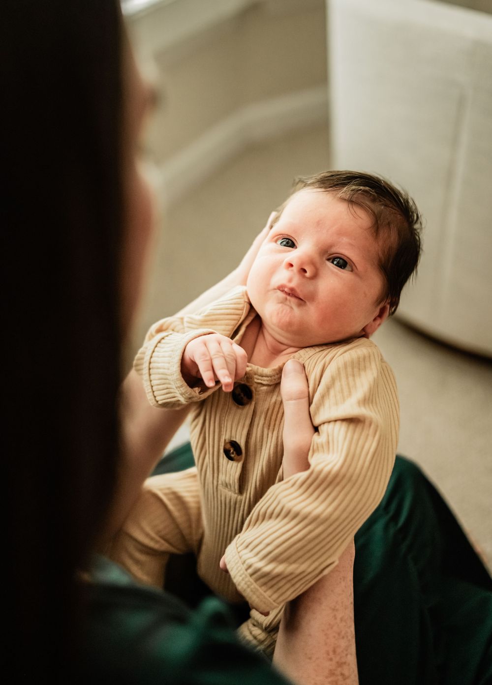 Baby in beige buttoned romper looking up while sitting on green fabric.