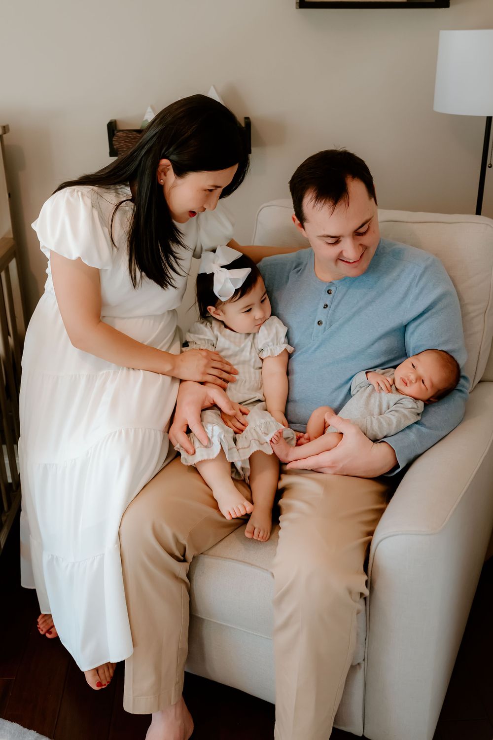 A family sits together on a white couch, with a small child and baby wearing white clothing in an indoor setting.