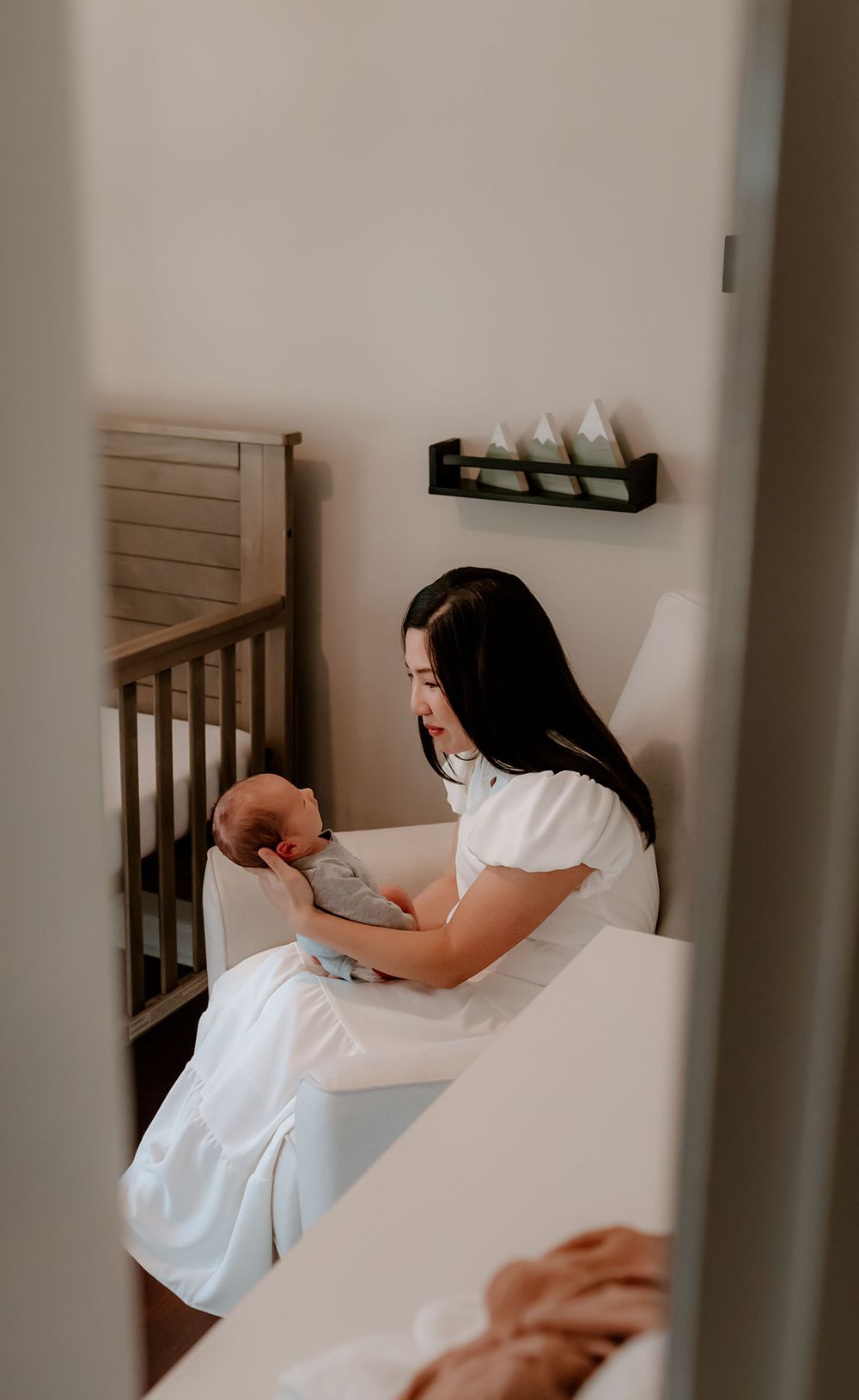 Woman in white dress sitting on stairs with baby, viewed through doorway in soft, moody lighting.