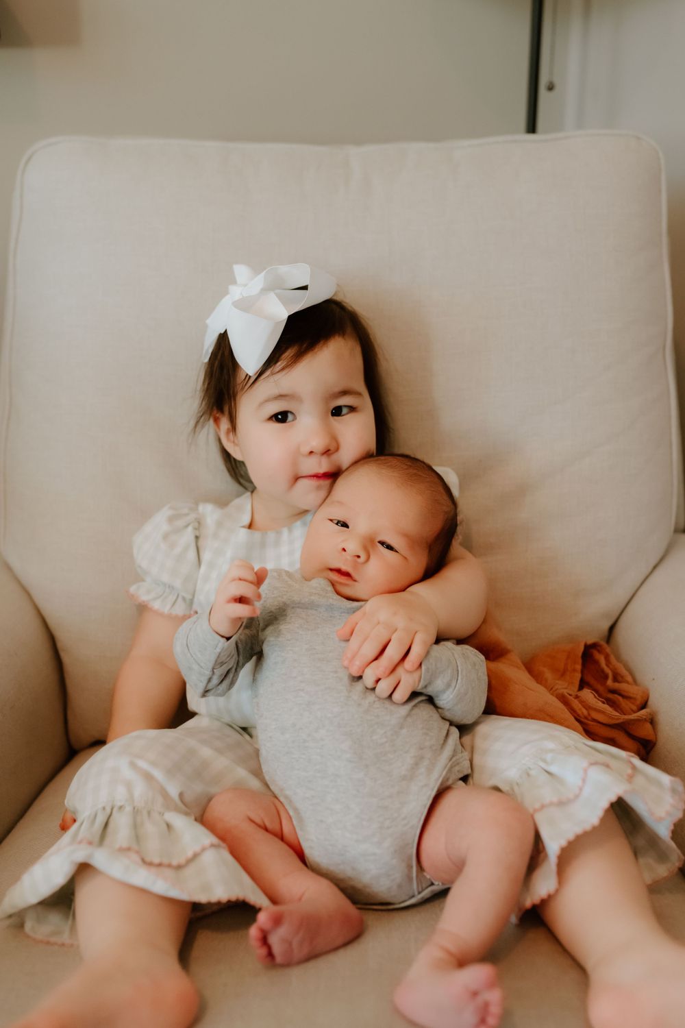 Two children sit together on a beige couch, with the older one wearing a white bow while holding a younger sibling.