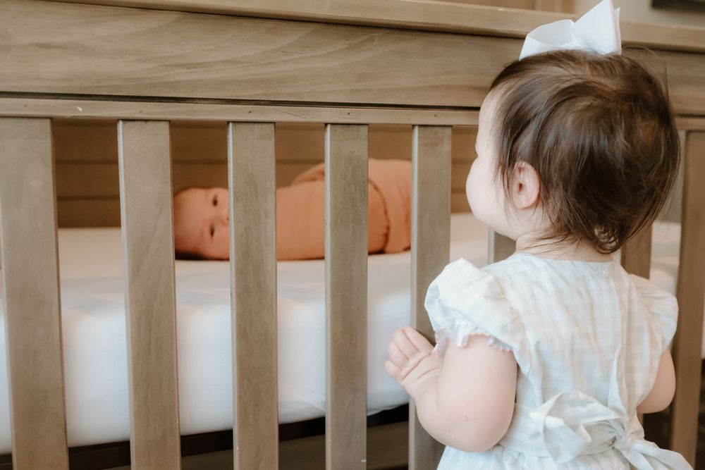 A small toddler in a white dress stands at a wooden crib, looking in at orange stuffed toys.