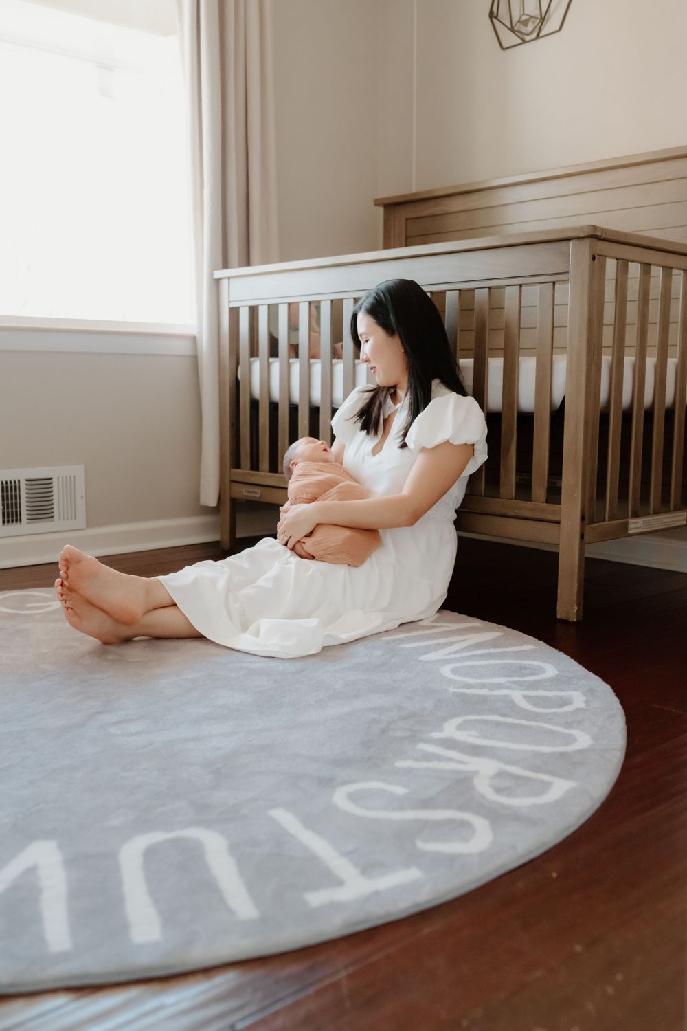 A woman in a white dress sits on a round gray rug near a wooden crib in a minimalist nursery.