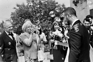 Black and white wedding photo of guests celebrating outdoors, woman photographing couple with camera, bubbles floating in air.