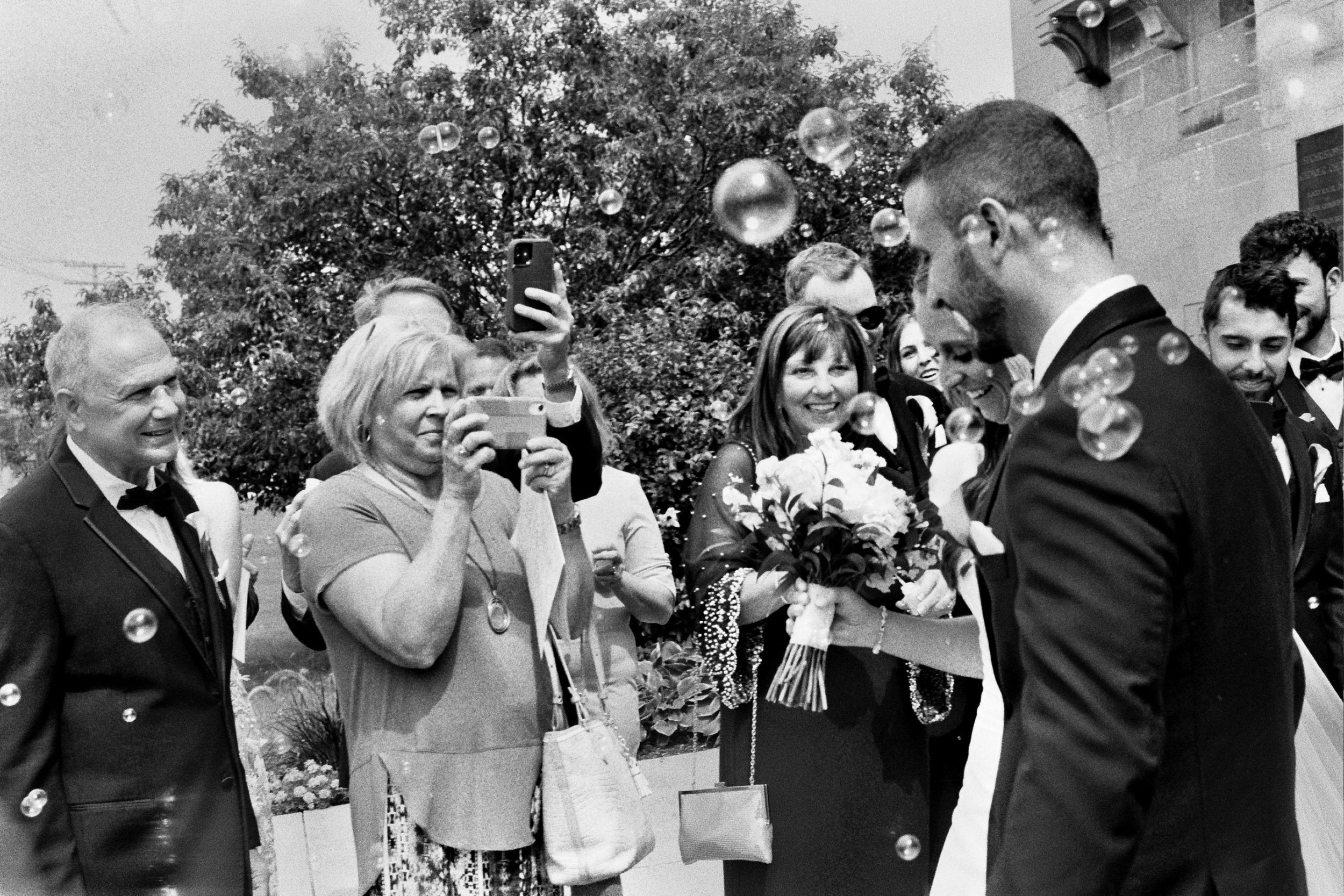 Black and white wedding photo of guests celebrating outdoors, woman photographing couple with camera, bubbles floating in air.