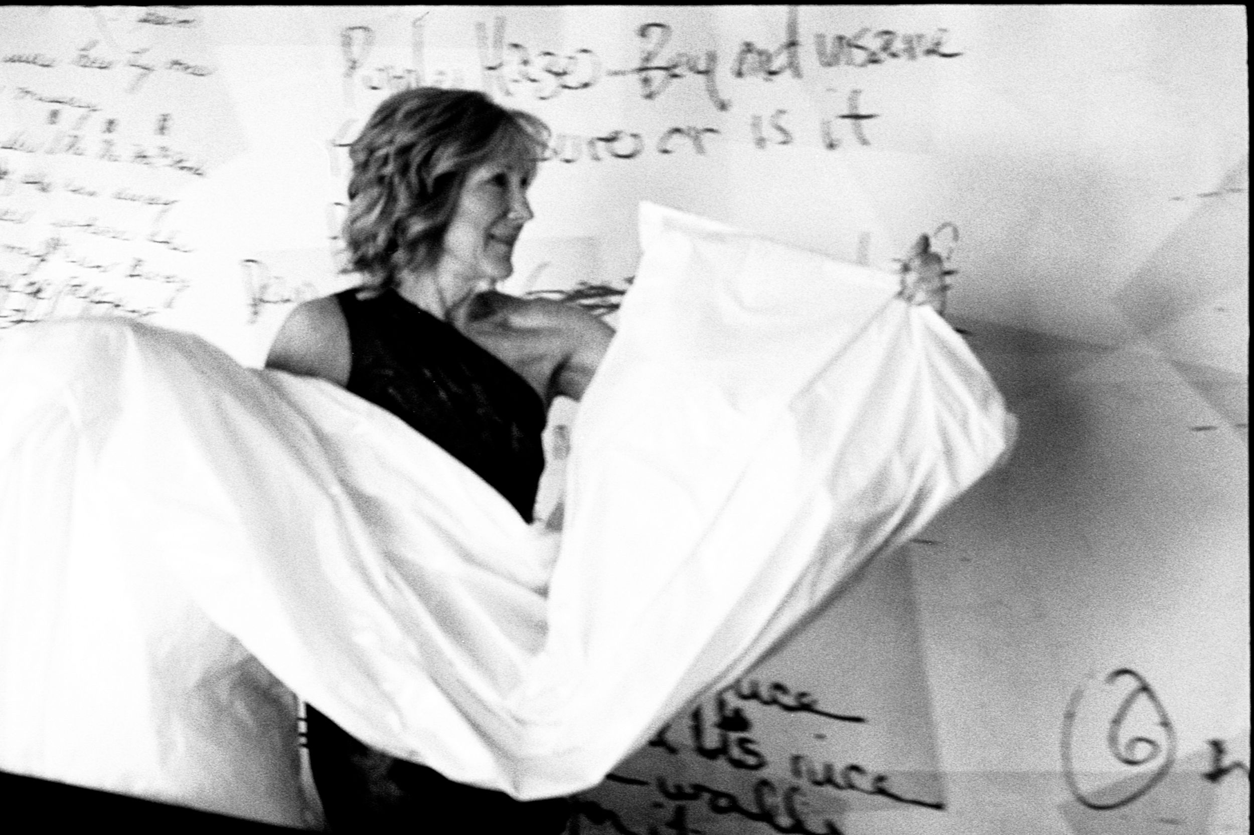 Black and white photo of a woman holding white fabric near a wall covered in handwritten text.