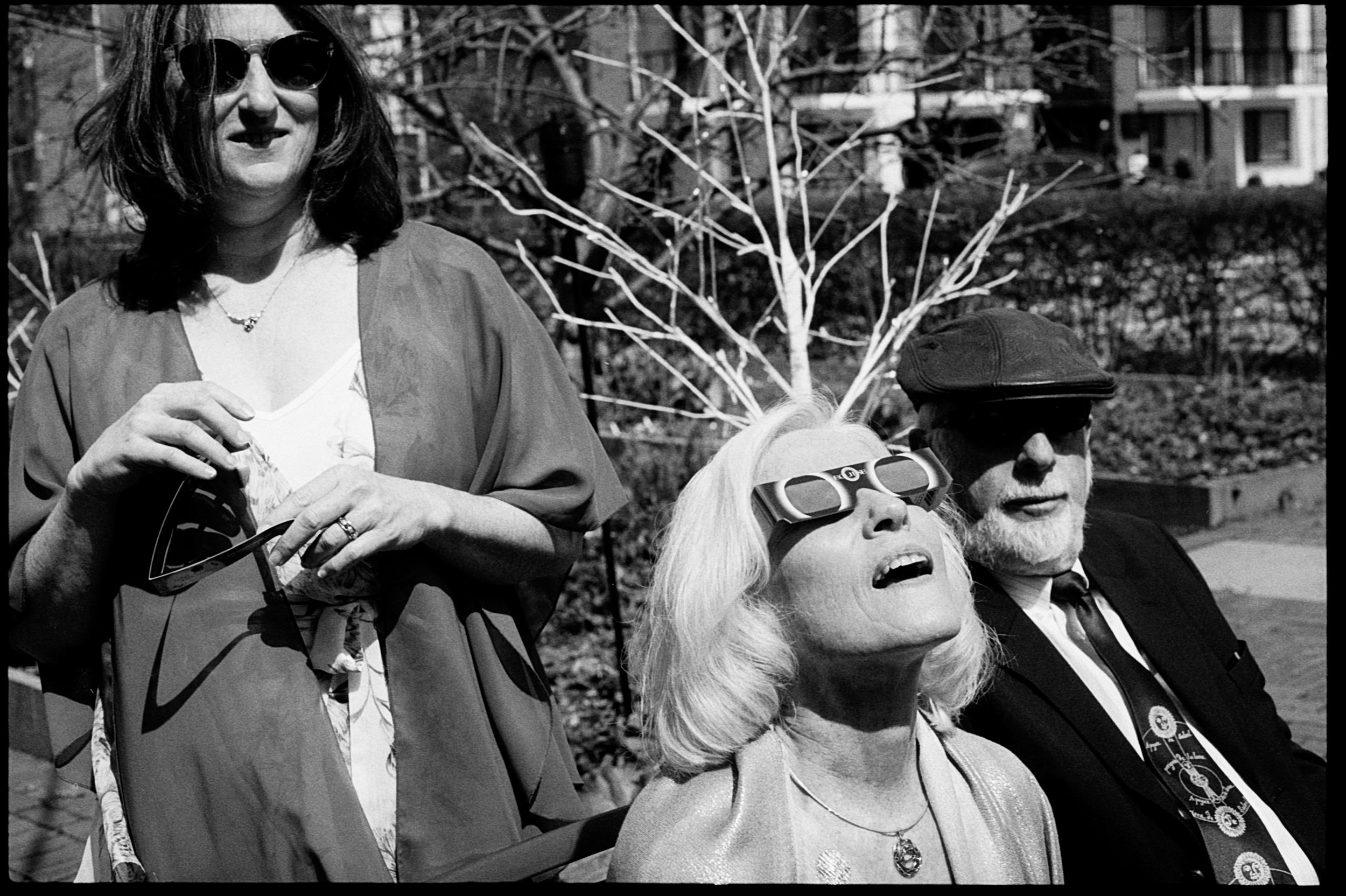 Black and white street photo of three people outdoors, woman in center wearing novelty sunglasses looking up.