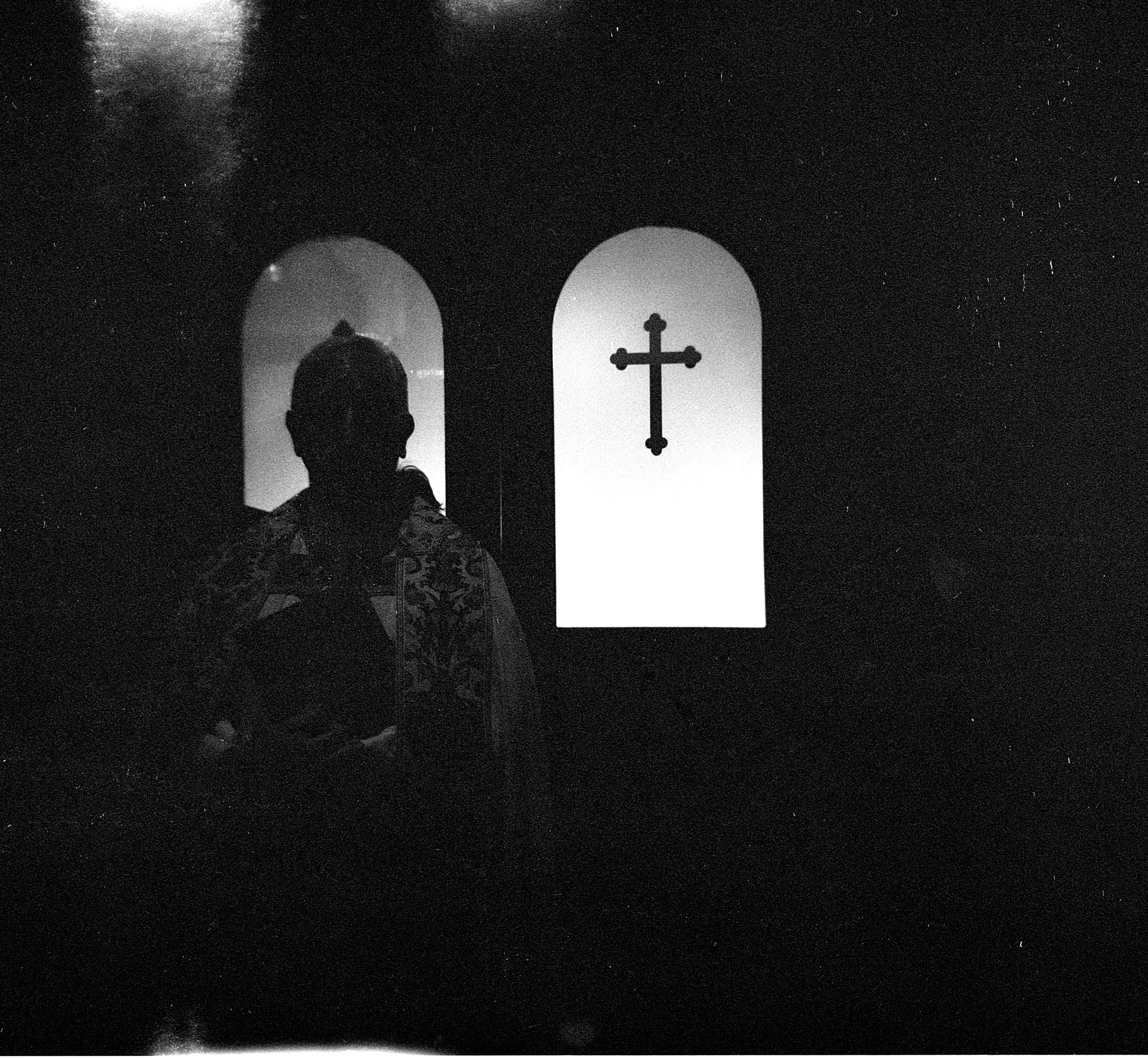 Dramatic black and white photo of a silhouetted figure beside an arched window with a cross.