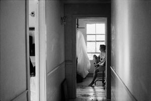 Black and white photo of a person sitting near a wedding dress hanging by a window.