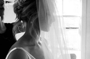 Black and white photo of a bride wearing a veil, viewed from behind near a bright window.