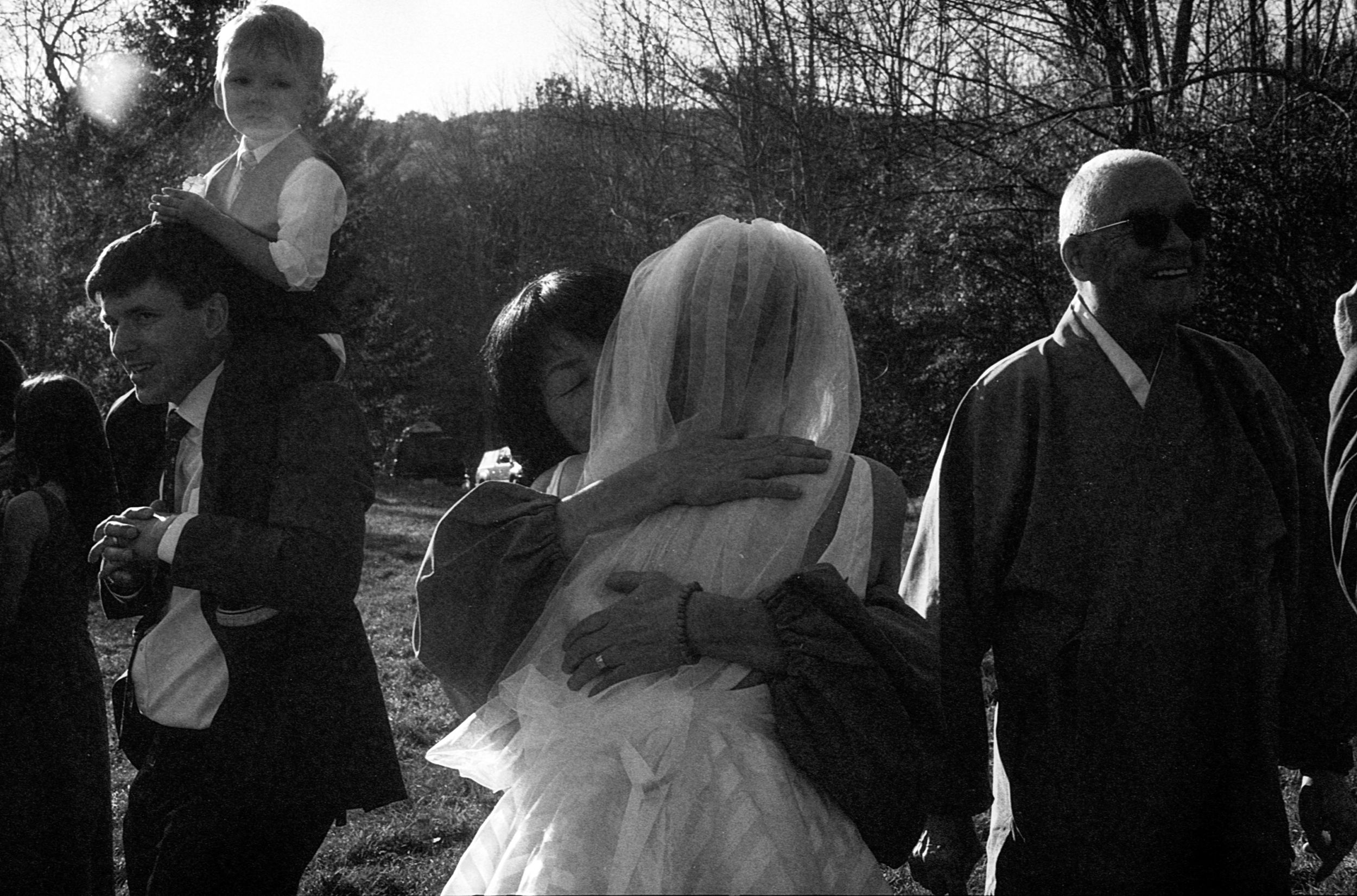 Black and white photo of a bride hugging a guest at an outdoor wedding reception with other attendees nearby.