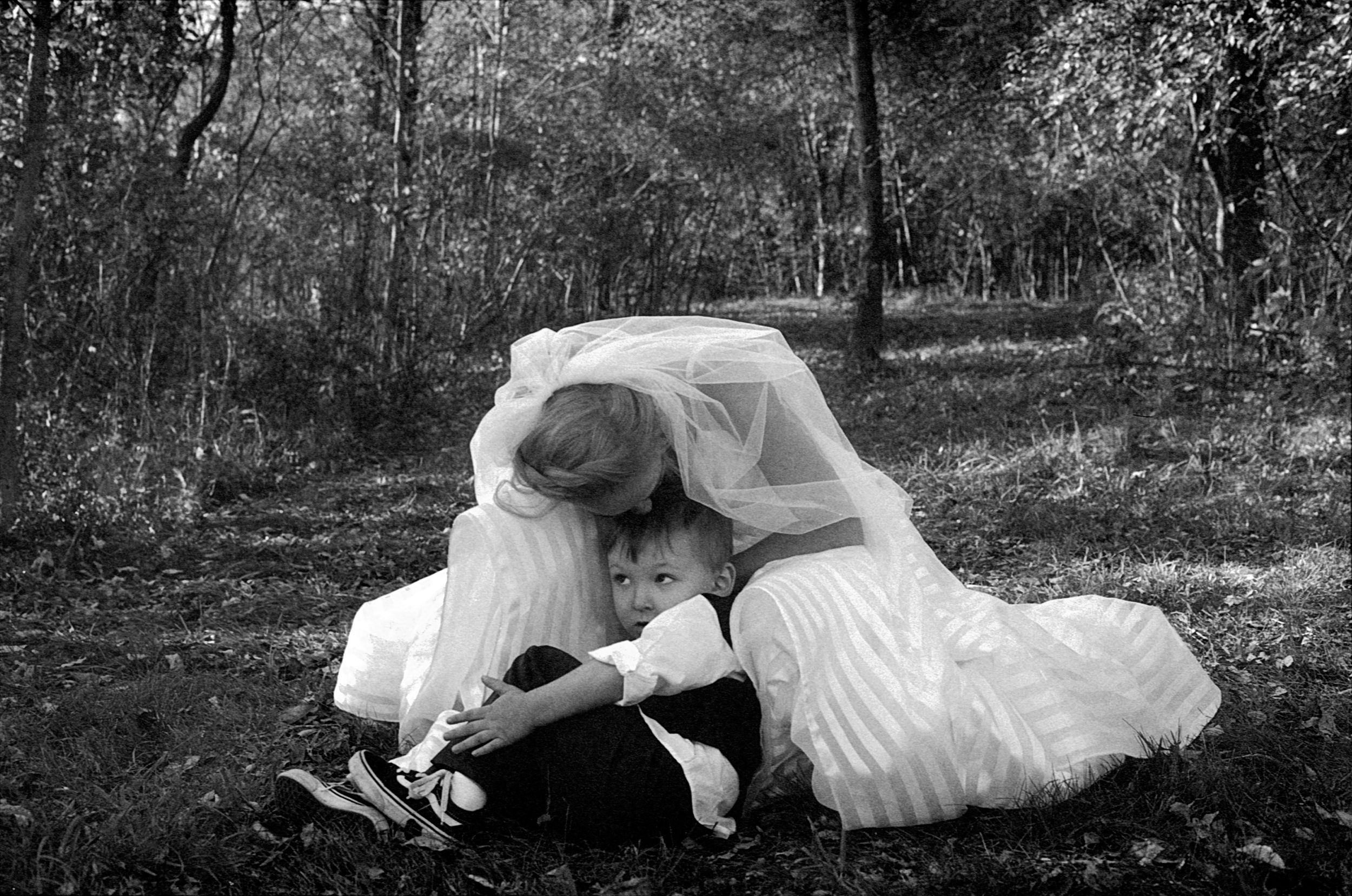 Black and white photo of a bride in white gown sitting on grass hugging a young child in a forest setting.