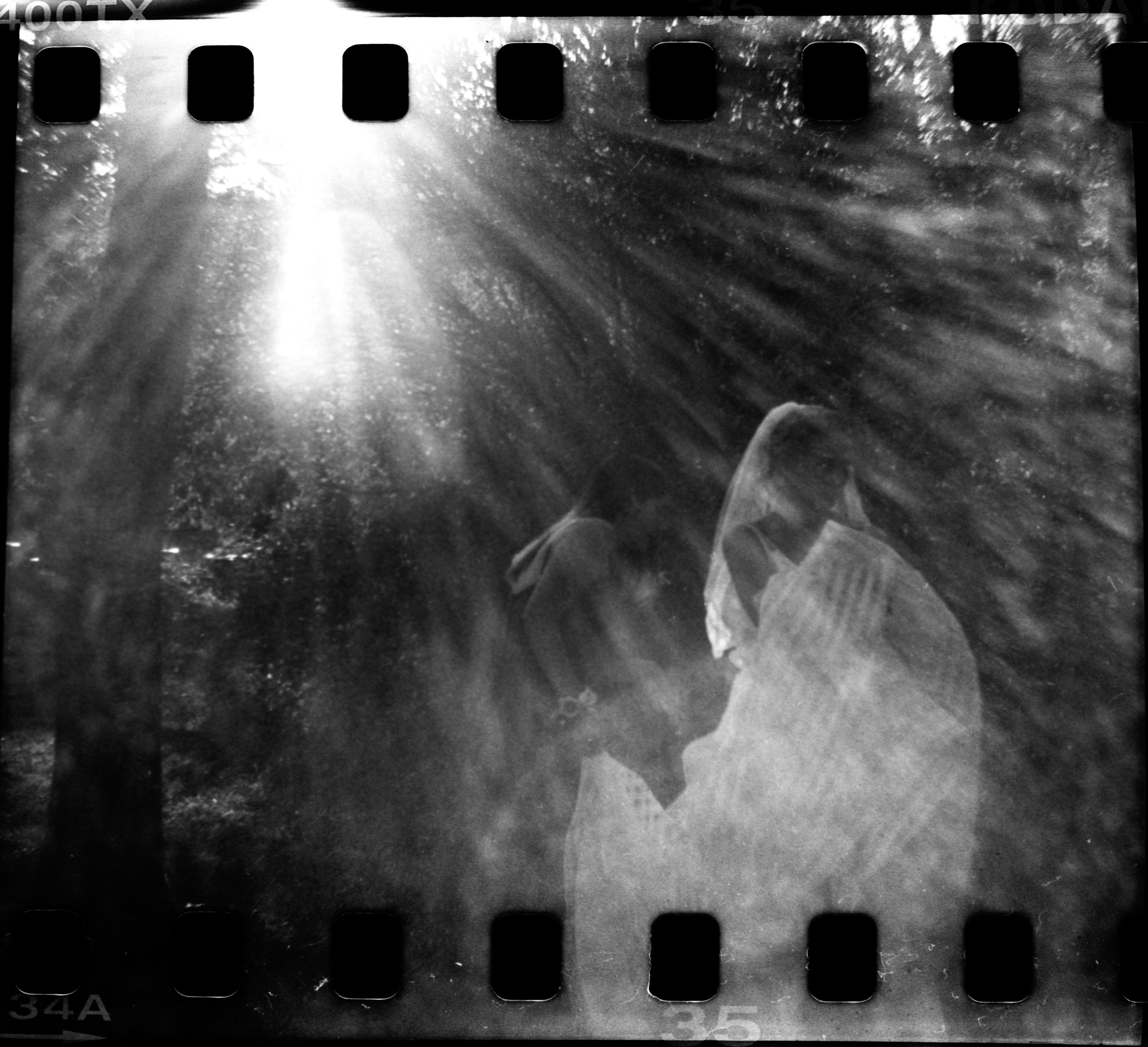 Dreamy black and white film photo of couple kissing with light rays in forest.