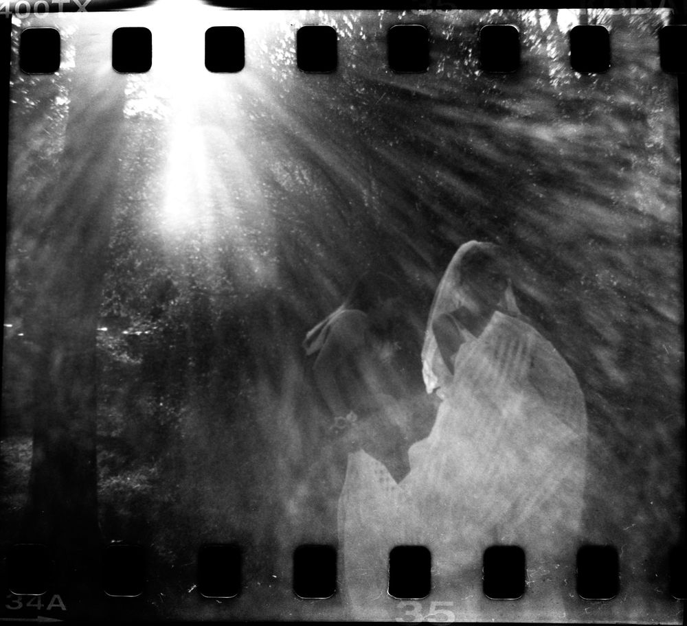 Dreamy black and white film photo of couple kissing with light rays in forest.