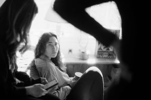 Black and white photo of a young woman with long hair sitting thoughtfully indoors, surrounded by people in a cozy room.