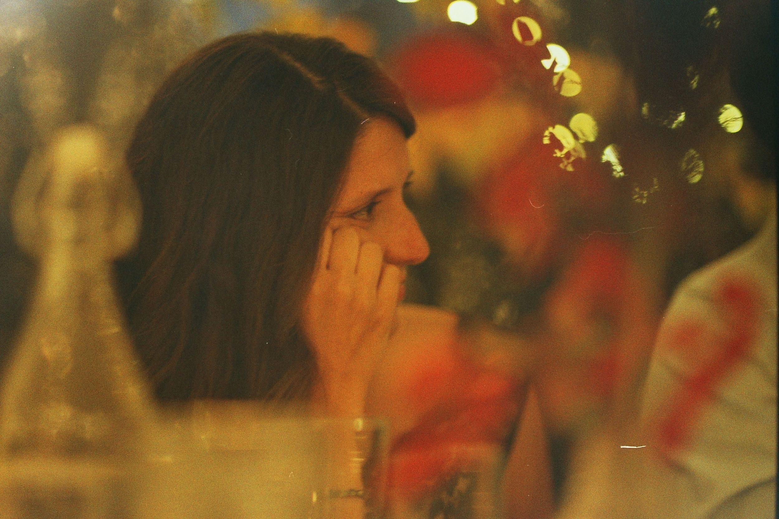 A pensive woman rests her hand on her face at a dimly lit gathering with bokeh lights in the background.