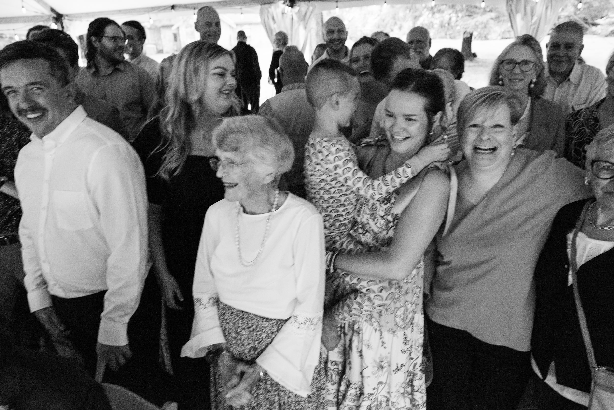 Black and white photo of multigenerational family laughing and embracing at an outdoor celebration.