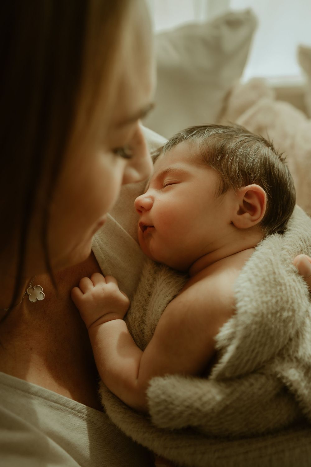 A mother tenderly holds her sleeping newborn baby wrapped in a soft blanket in a warm, intimate setting.