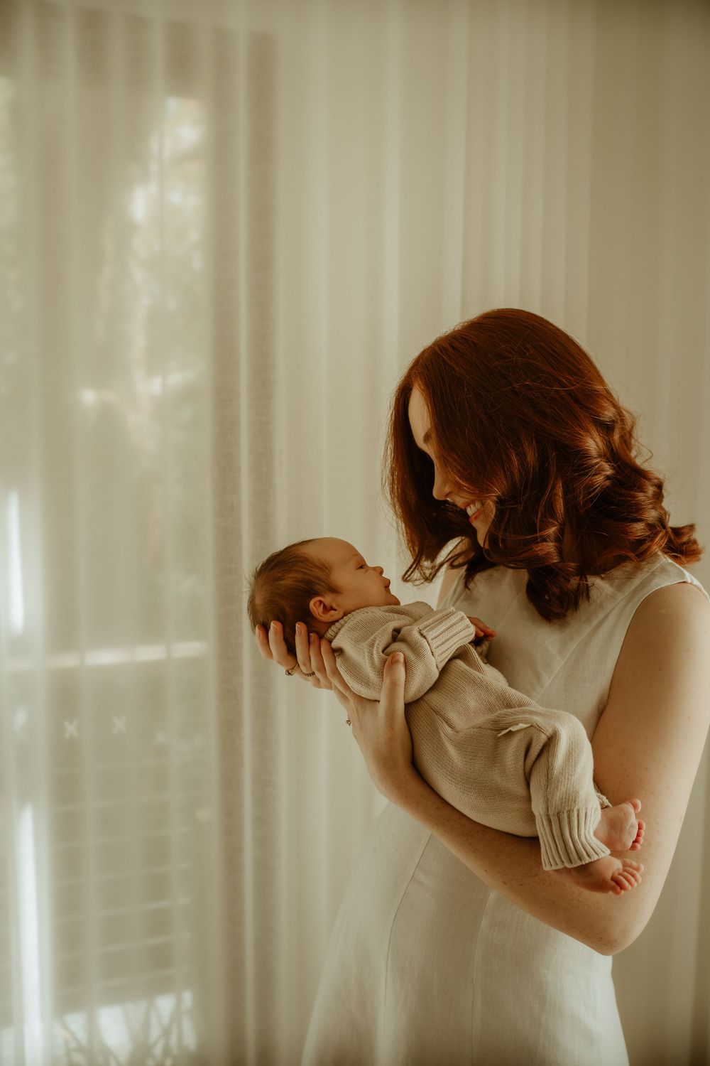 A tender moment between parent and newborn baby in soft window light, captured in warm sepia tones.