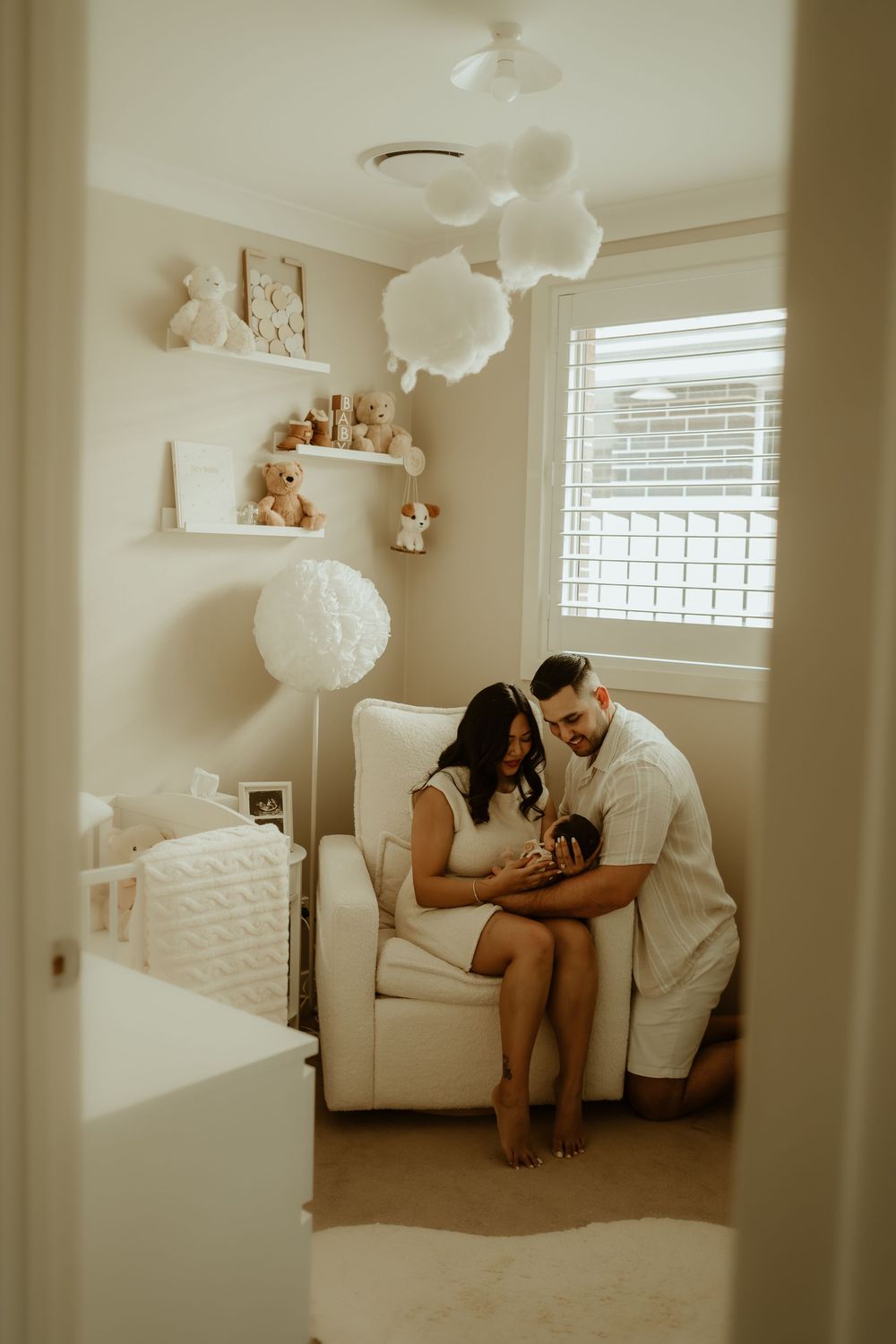 A couple sits together in a cozy nursery with white decor, cloud pendant lights, and floating shelves with stuffed animals.