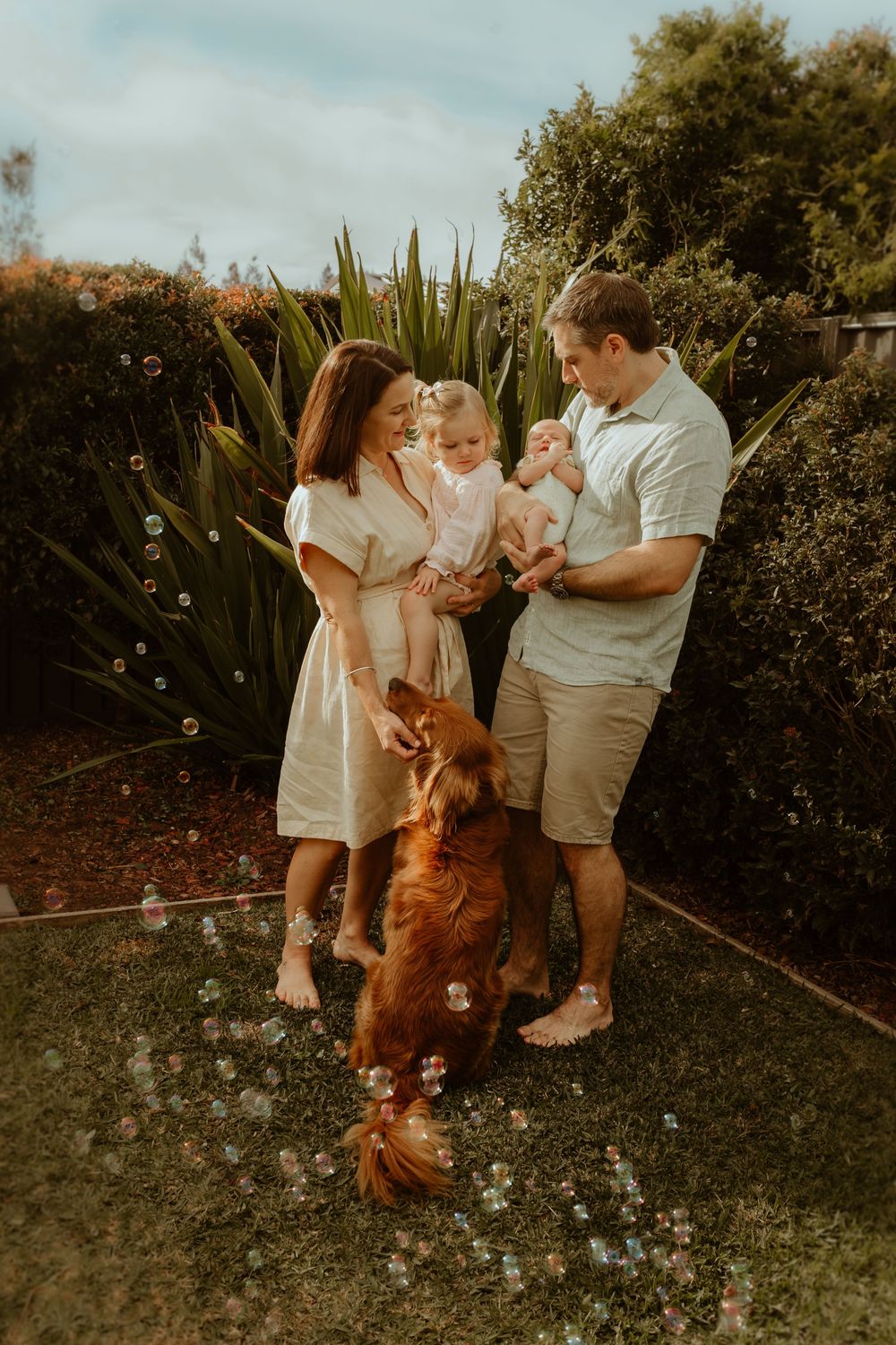A family stands together in a garden with their golden retriever dog while bubbles float through the warm sunlit air.