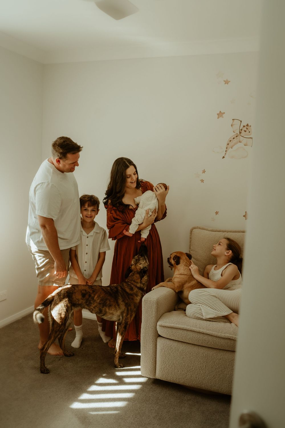 Family with newborn baby, two children, and two dogs gathered in a warmly lit nursery room.