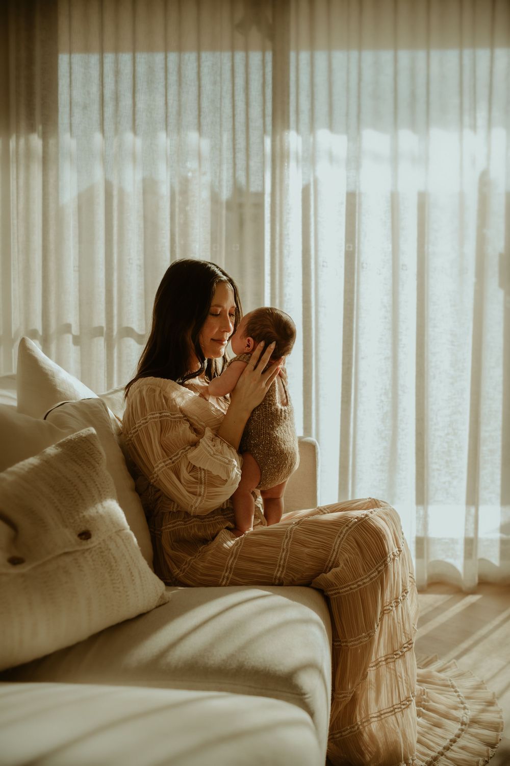 Mother and baby share tender moment on white couch in sunlit room with sheer curtains.