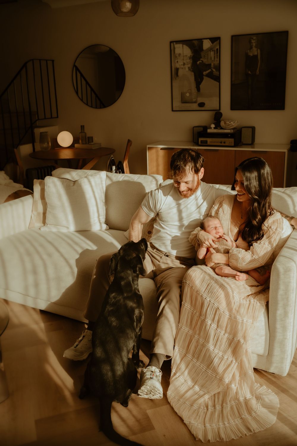 Romantic couple lounges on white couch with black dog in warm sepia-toned living room setting.