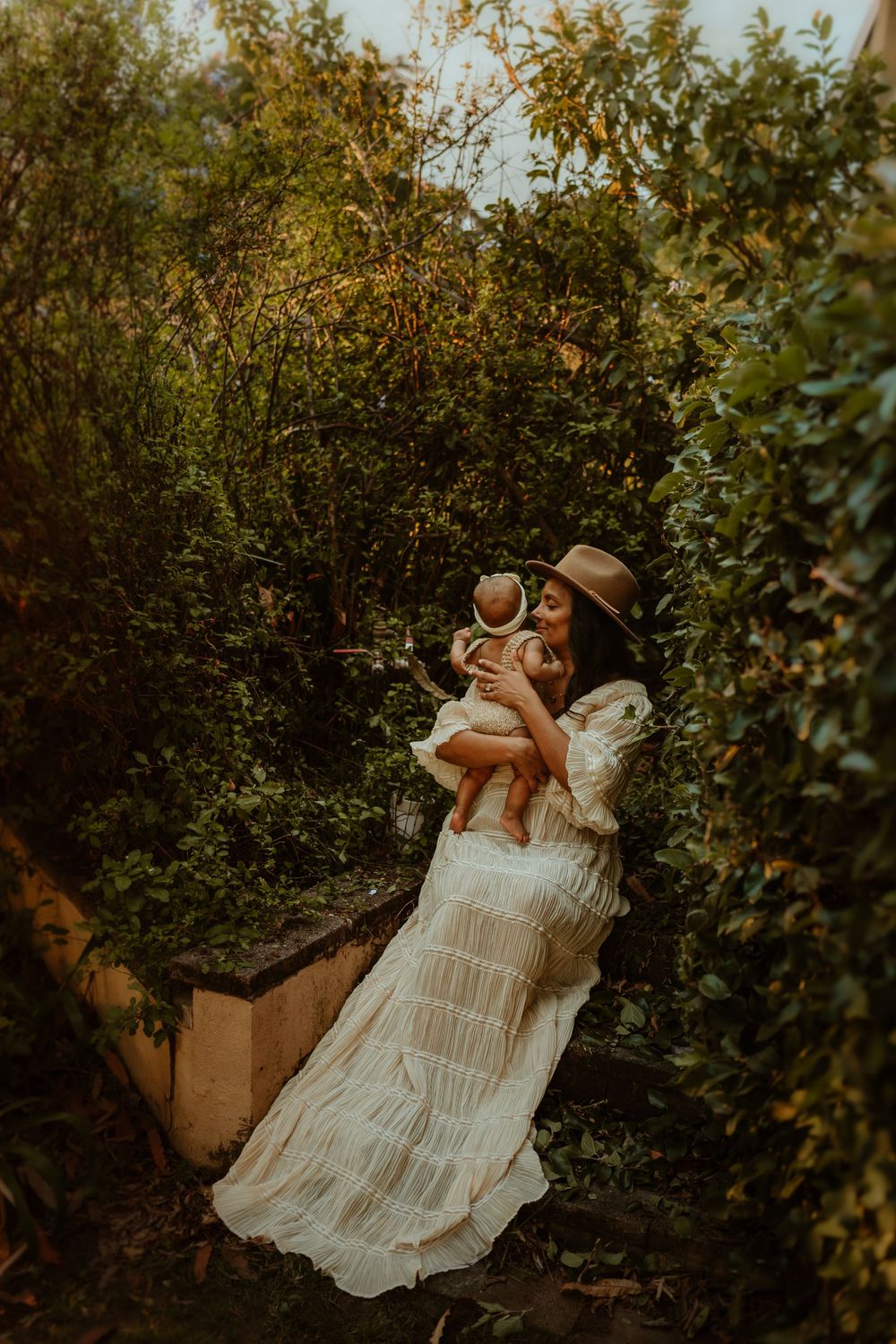 A woman in a white boho dress and hat holds her baby among lush green garden foliage in warm golden light.