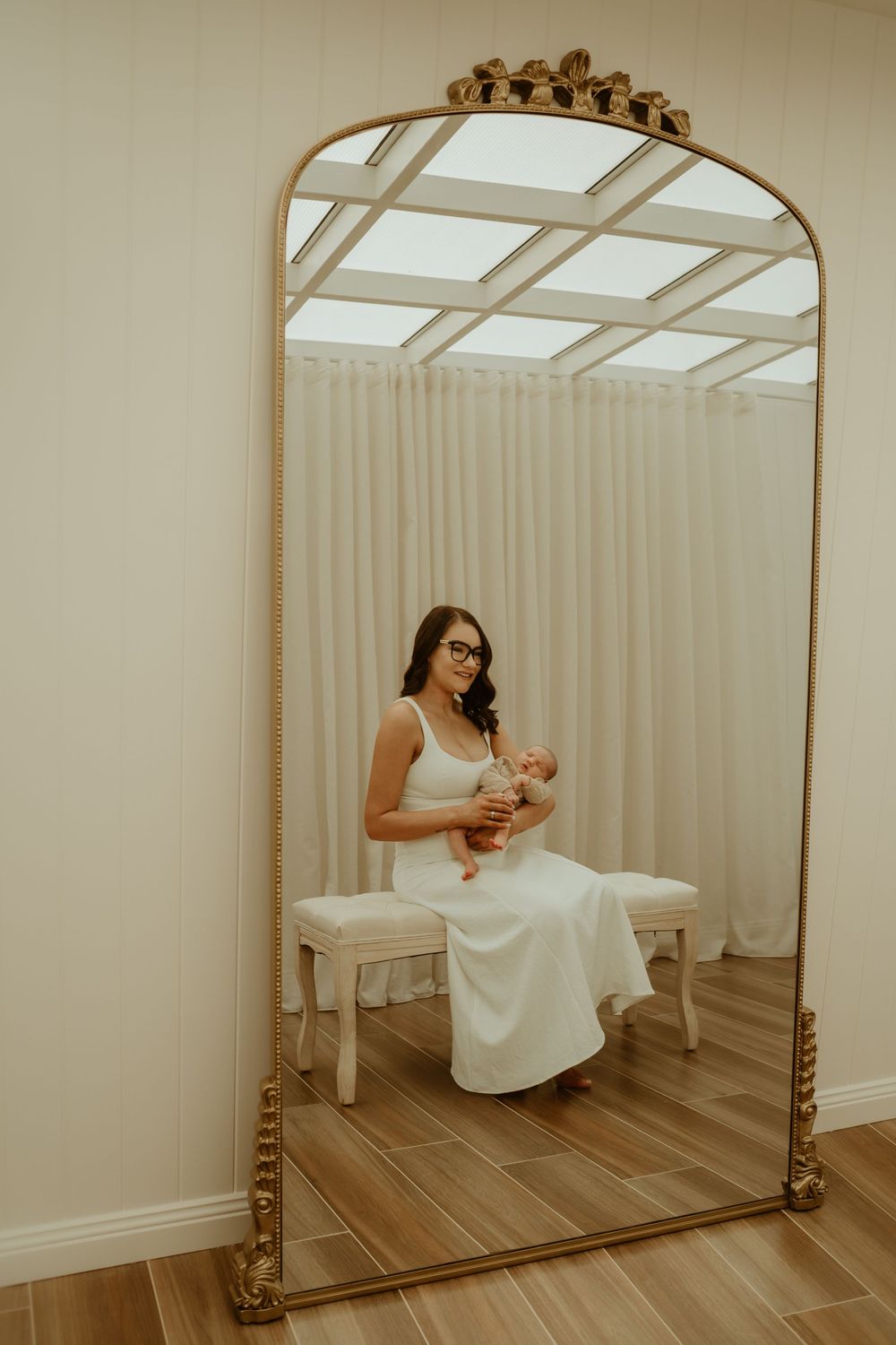 A woman in a white dress sits on a bench holding a newborn, reflected in an ornate gold arch mirror.