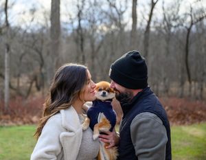 A couple holds and kisses their small Pomeranian puppy in a winter forest setting.