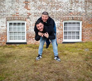 A person in a black jacket and jeans crouches on a grassy lawn in front of a brick building with white windows.