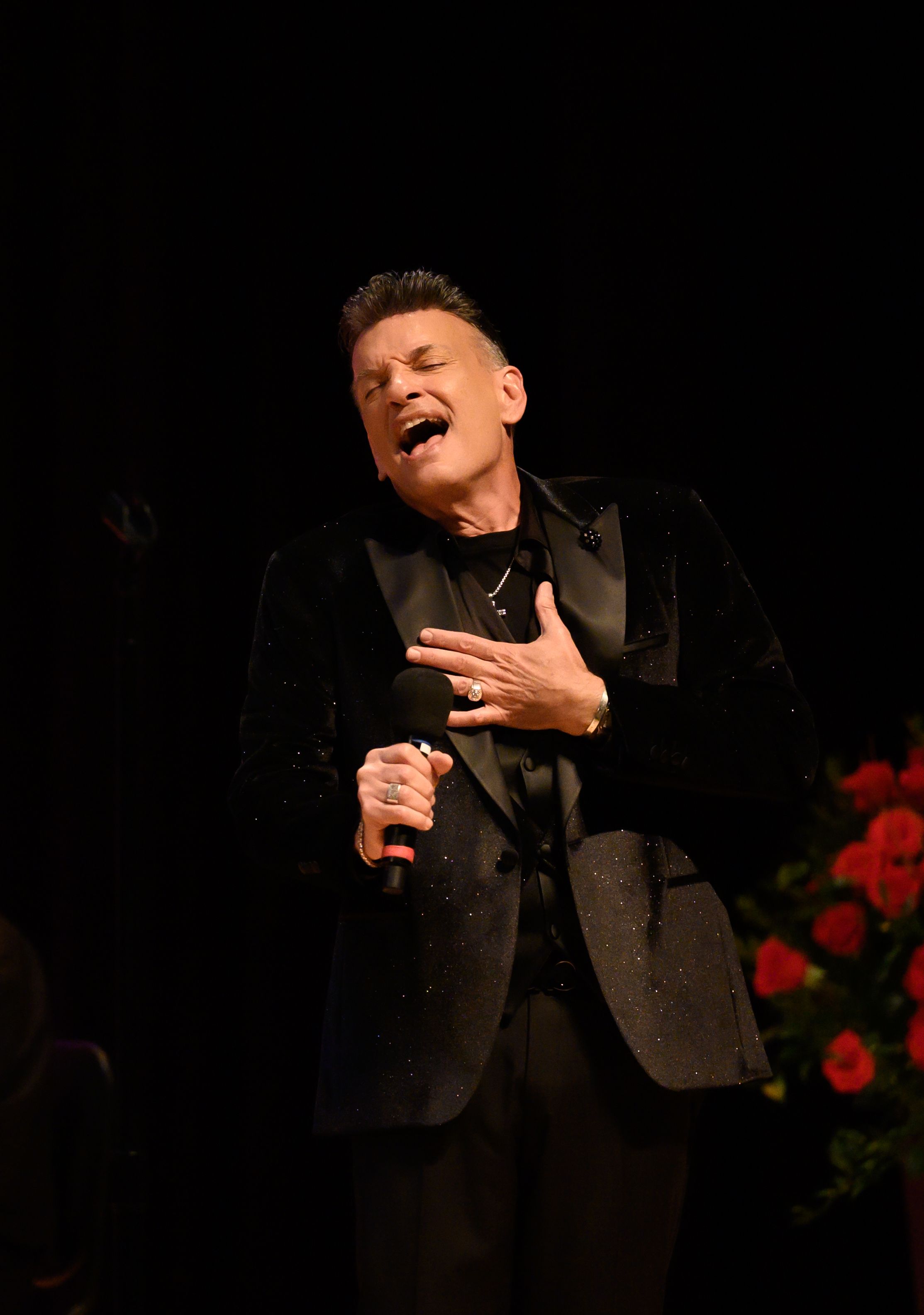 A performer in a dark suit sings passionately on stage during a dramatic performance in low lighting.