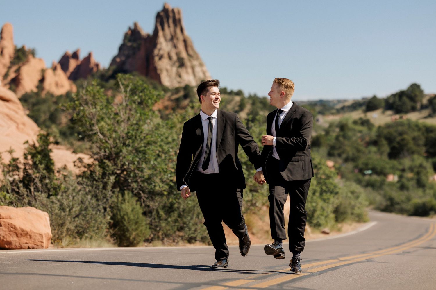 A groom and his husband joyfully run down a road with Colorado's iconic Garden of the Gods in the background