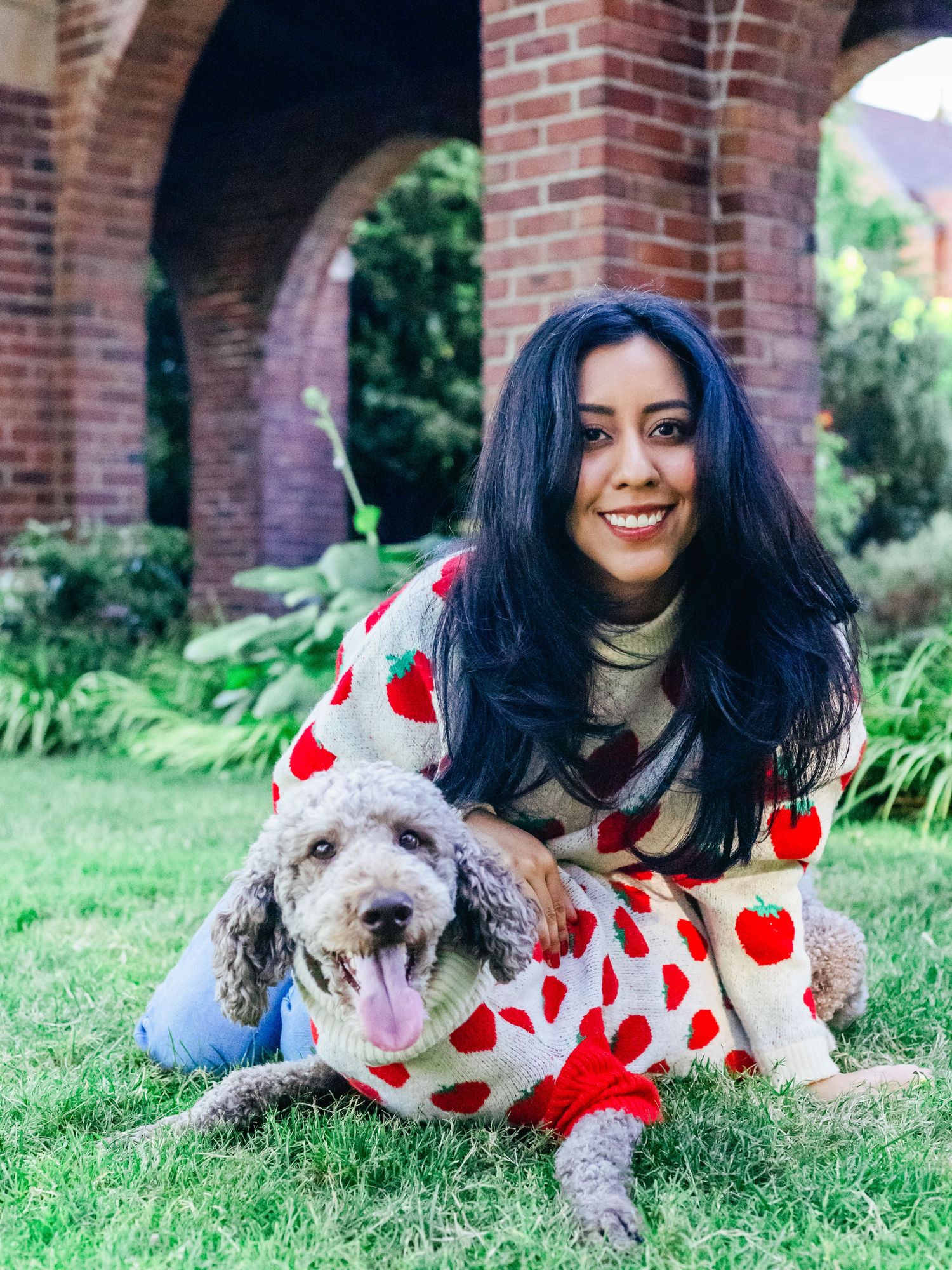 A smiling woman in a strawberry sweater hugs her dog, also in a matching sweater, on a green lawn with brick arches and plants in the back.