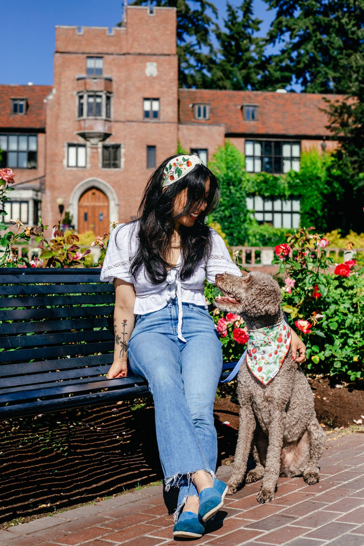 A woman in a white top and jeans sits on a bench, smiling at her fluffy dog in a bandana. They're in a garden with a brick building behind.
